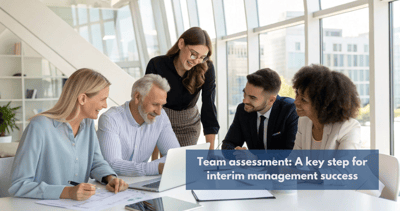 The image shows a group of five professionals gathered around a white conference table in a bright, modern office setting with large windows overlooking a cityscape. Four individuals are seated, while one woman stands slightly behind and between two of the seated people, leaning slightly forward as if engaged in the discussion.  On the table, there is a laptop open towards the group, along with some papers, pens, and a smartphone. The atmosphere appears collaborative and engaged, with several members of the group smiling and looking towards the laptop screen or each other.