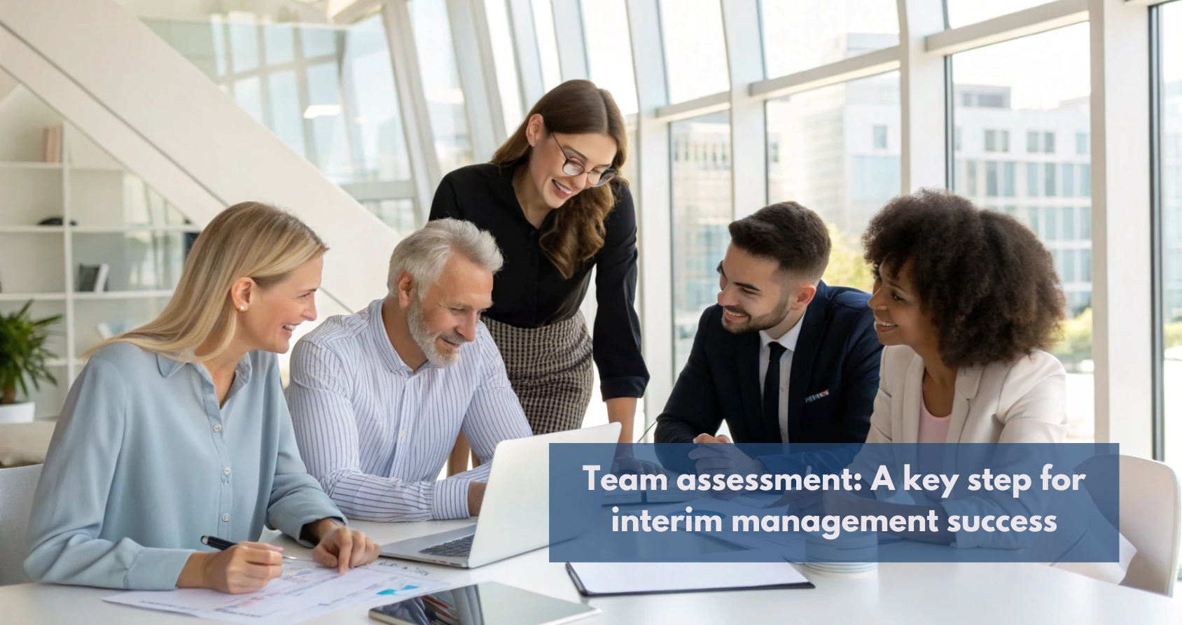 The image shows a group of five professionals gathered around a white conference table in a bright, modern office setting with large windows overlooking a cityscape. Four individuals are seated, while one woman stands slightly behind and between two of the seated people, leaning slightly forward as if engaged in the discussion.  On the table, there is a laptop open towards the group, along with some papers, pens, and a smartphone. The atmosphere appears collaborative and engaged, with several members of the group smiling and looking towards the laptop screen or each other.