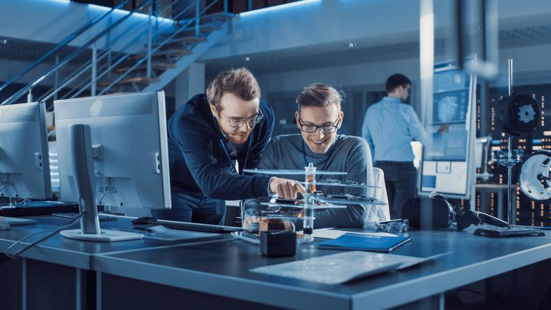Dos ingenieros revisan un componente electrónico en un laboratorio de oficina técnica mientras un tercer profesional trabaja al fondo.